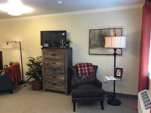 A cozy living room corner with an armchair, lamp, and a wooden dresser topped with a television.