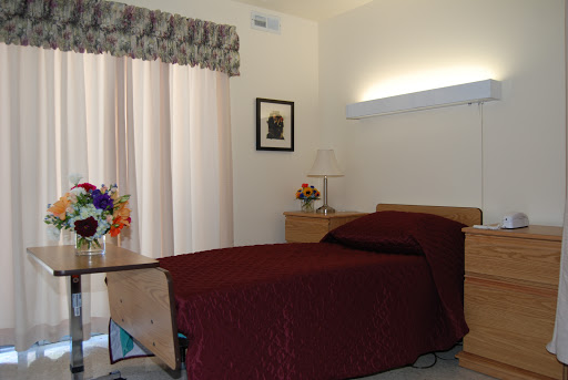 A cozy bedroom featuring a maroon bedspread, curtains, and a small flower arrangement.