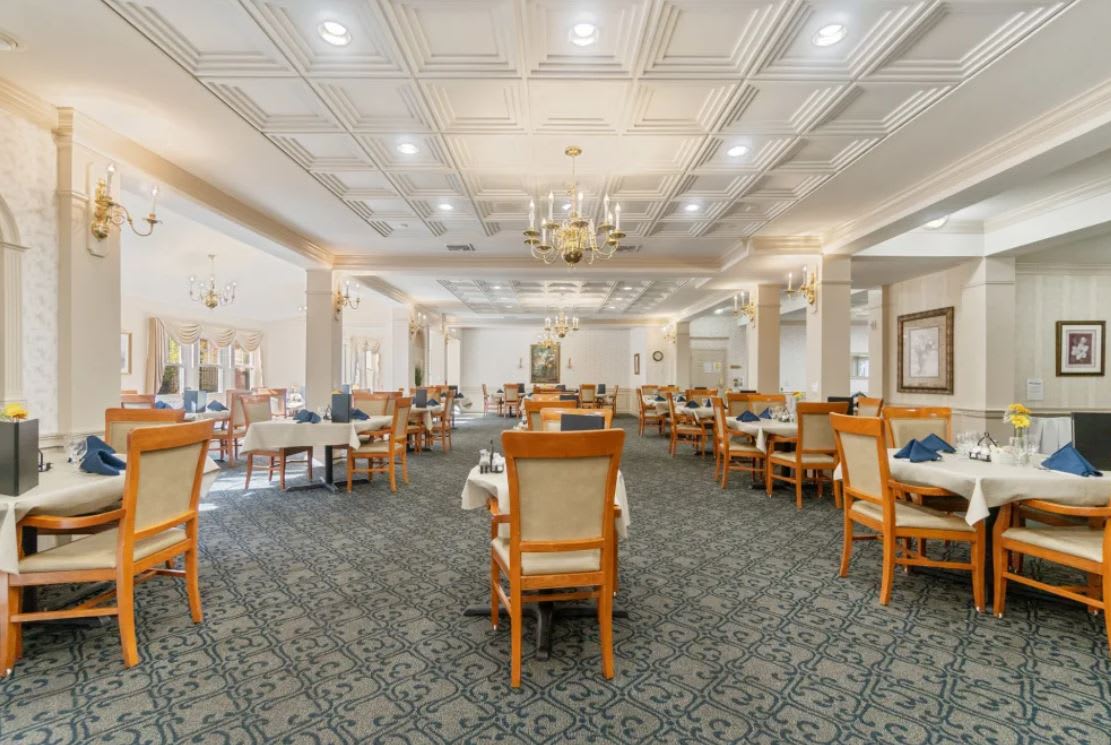 A spacious dining room with elegant chandeliers, neatly arranged tables and chairs, and a carpeted floor.