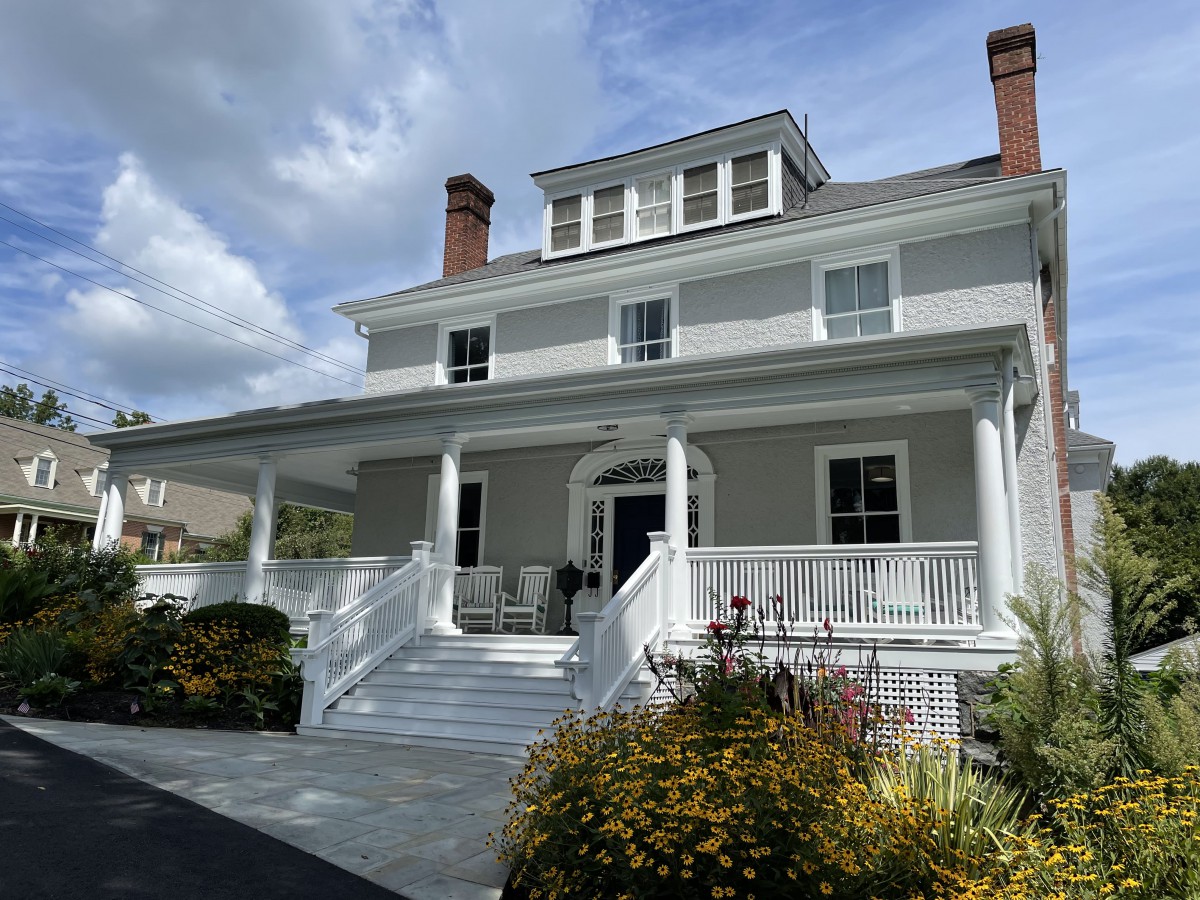 A large white house with a spacious porch and surrounding garden under a partly cloudy sky.