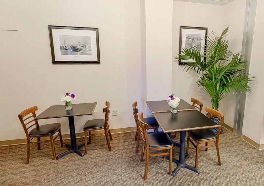 A cozy cafe interior with tables and chairs arranged neatly, adorned with framed artwork and a potted plant.