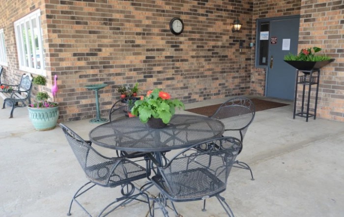 A cozy patio setting with an iron table and chairs against a brick wall, featuring a potted plant and a wall-mounted clock.