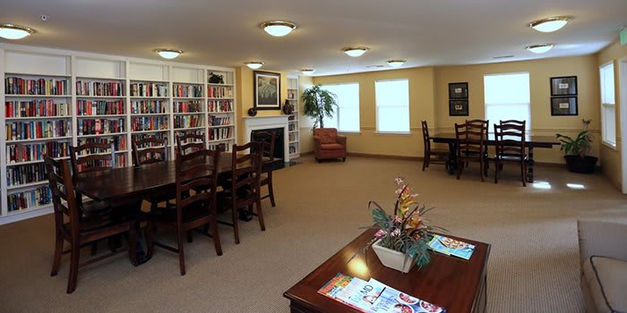 A cozy library with bookshelves, tables, and chairs, featuring a fireplace and indoor plant.