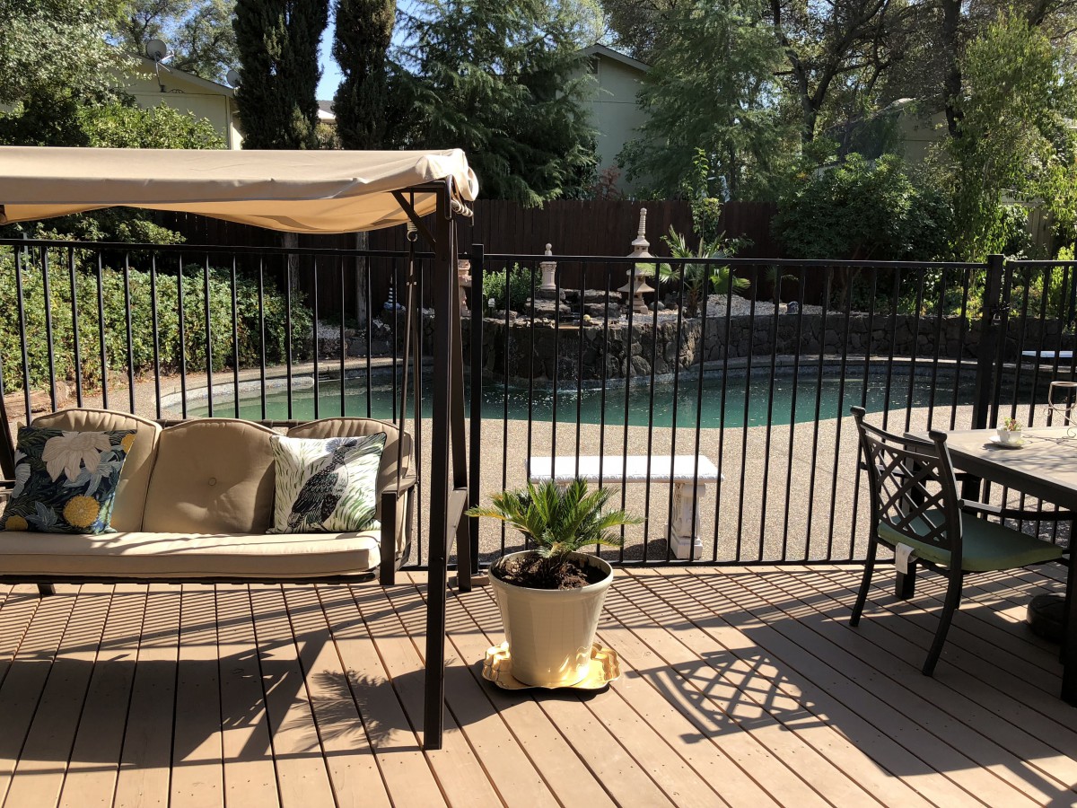 A sunny patio with a swing and potted plants next to a fenced swimming pool.