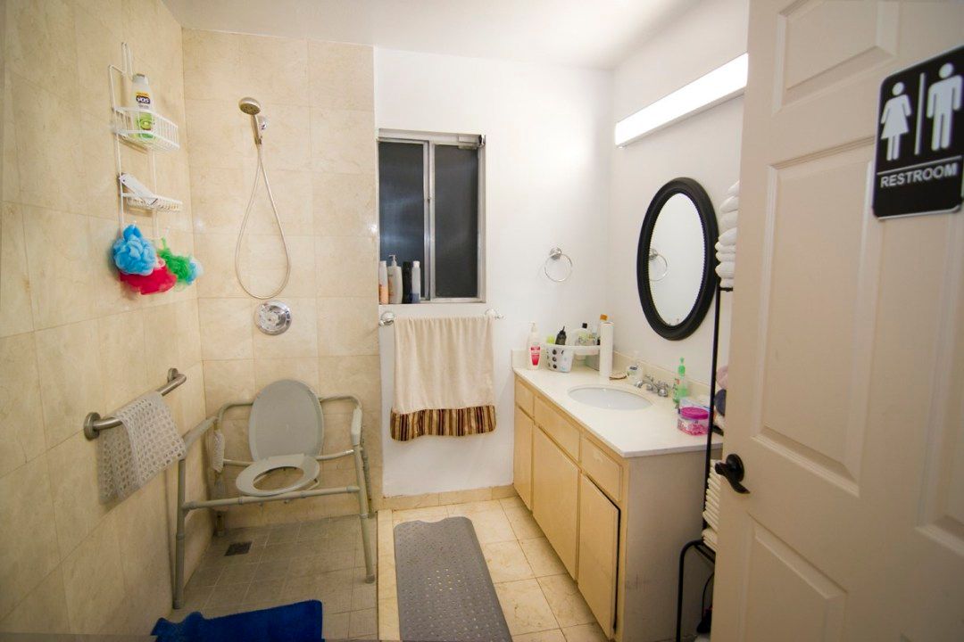 A well-lit bathroom featuring a shower area with safety chair, a vanity with a round mirror and sink, and neatly arranged toiletries.