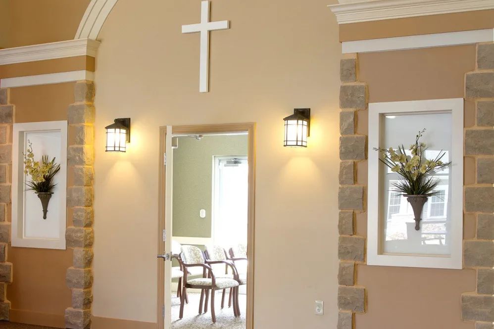 A serene room interior with a cross above the doorway, leading to a space with chairs and warm lighting.