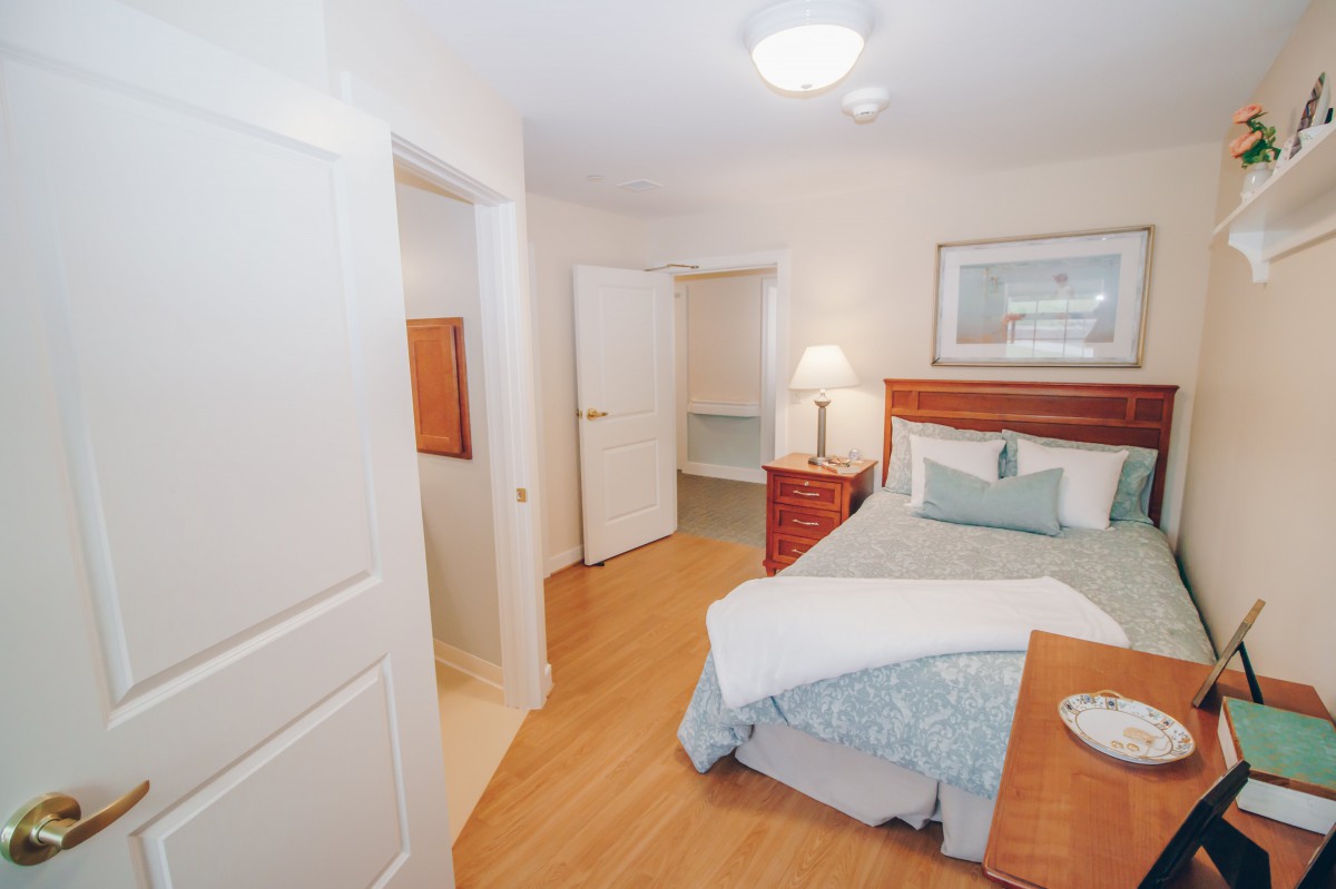 A cozy bedroom with wooden flooring, featuring a bed, a bedside lamp, and artwork on the wall.