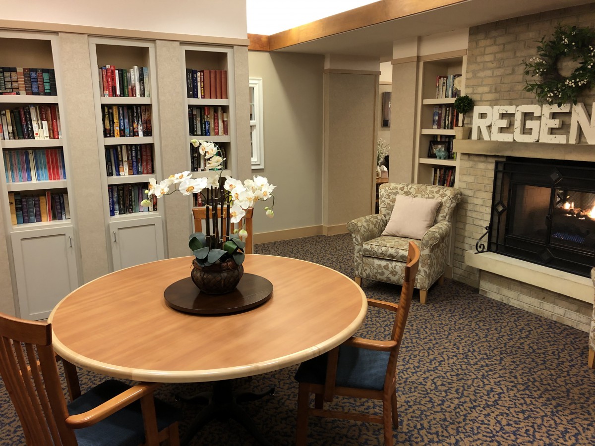 A cozy library reading room with a wooden table, surrounded by shelves filled with books and a fireplace.