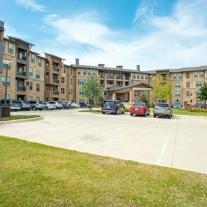 A residential apartment complex with a parking lot in the foreground under a blue sky.