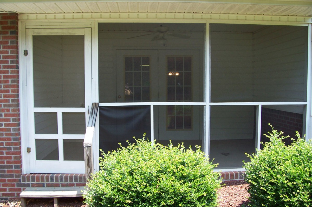 A screened porch with a damaged section, surrounded by bushes.