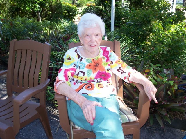 An elderly woman sitting outdoors in a garden, wearing a colorful shirt and enjoying a sunny day.