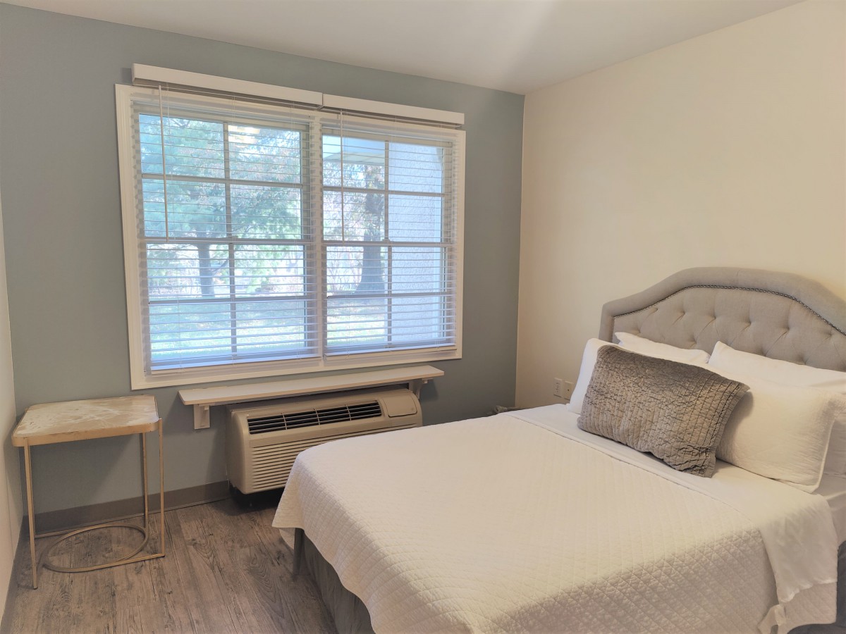 A modern and minimalist bedroom with natural light streaming through a large window, highlighting the neutral color scheme and simple furnishings.