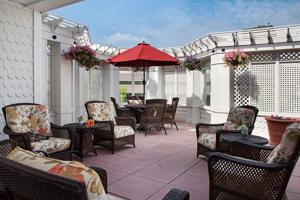 A cozy outdoor patio with comfortable furniture and a red umbrella under a pergola.