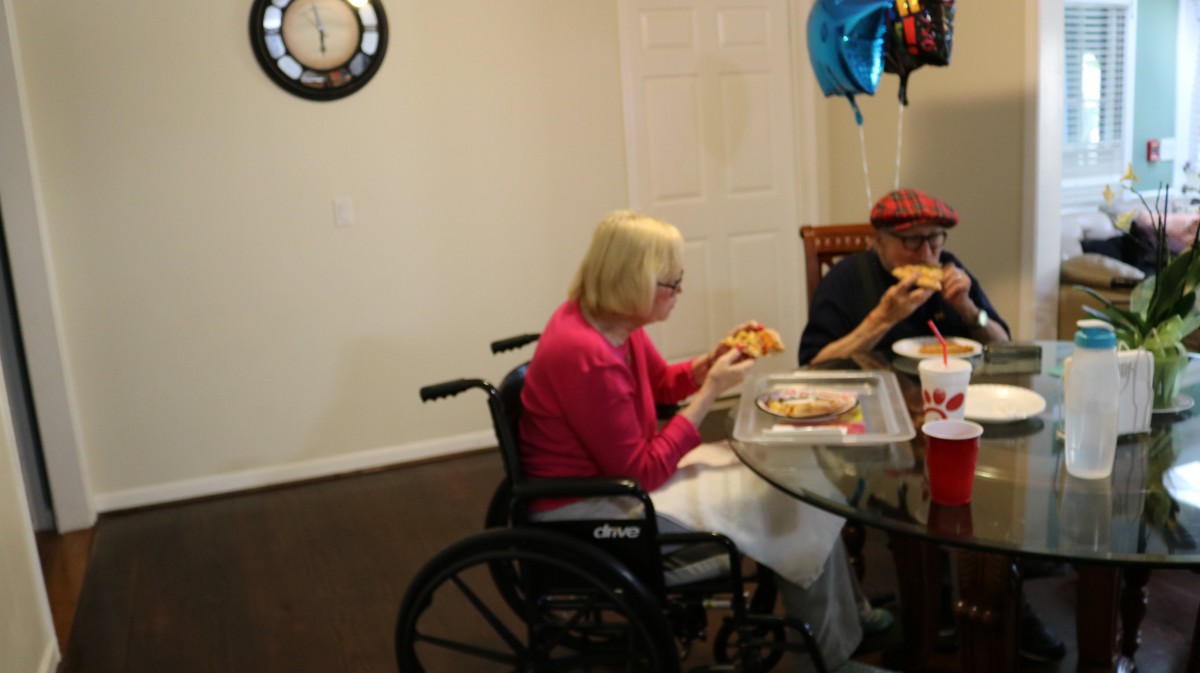 An elderly person in a wheelchair enjoying pizza with another person at a dining table, decorated with balloons.