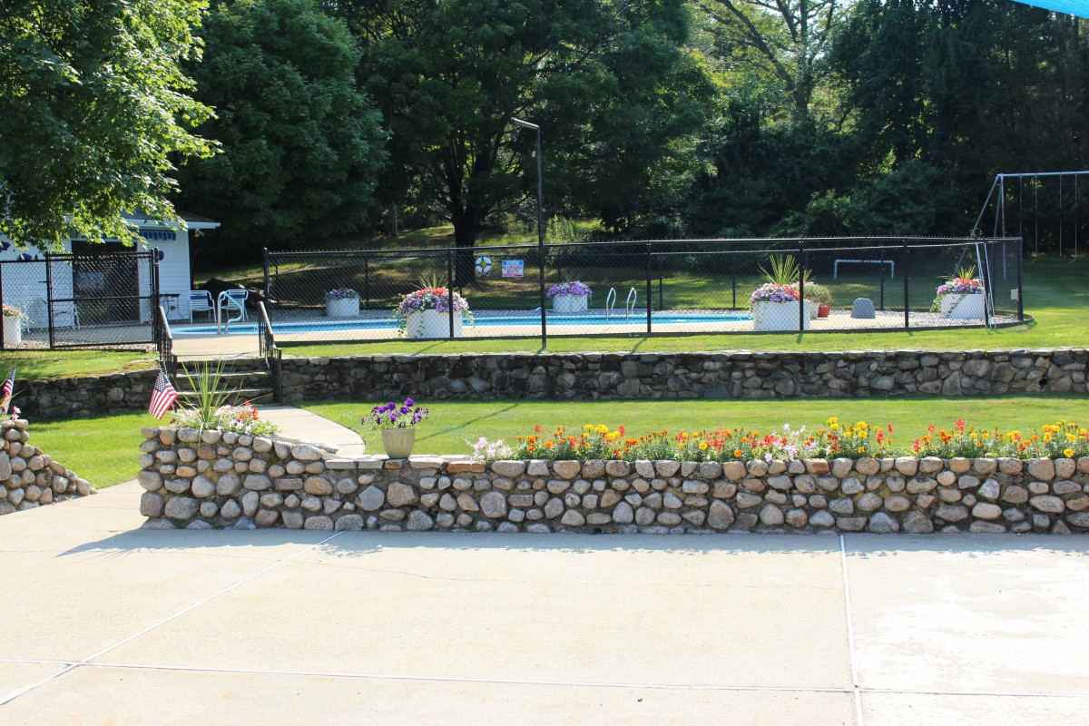 A fenced swimming pool area surrounded by greenery and a stone wall.