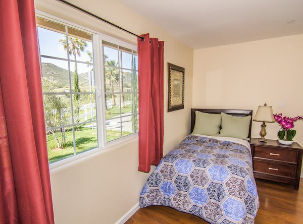 A cozy bedroom with a single bed by the window, featuring red curtains and a wooden nightstand.