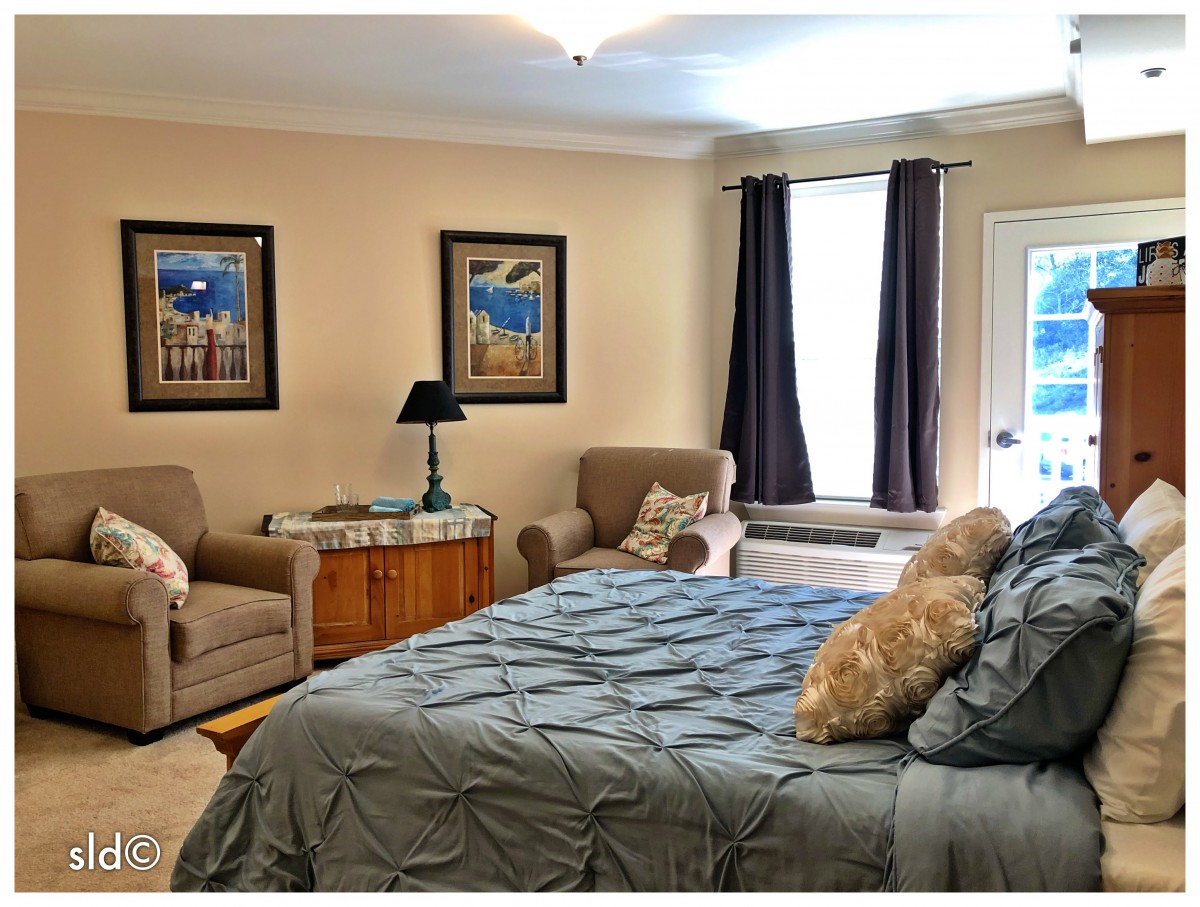 A cozy bedroom featuring a large bed with a blue comforter, two armchairs, decorative wall art, and a window with curtains.