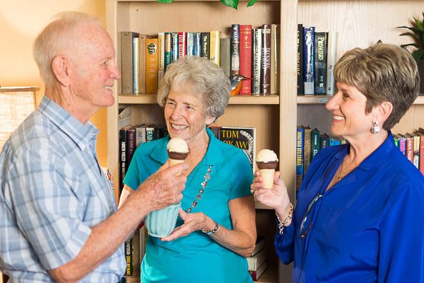 Three elderly people enjoying ice cream together in front of a bookshelf.