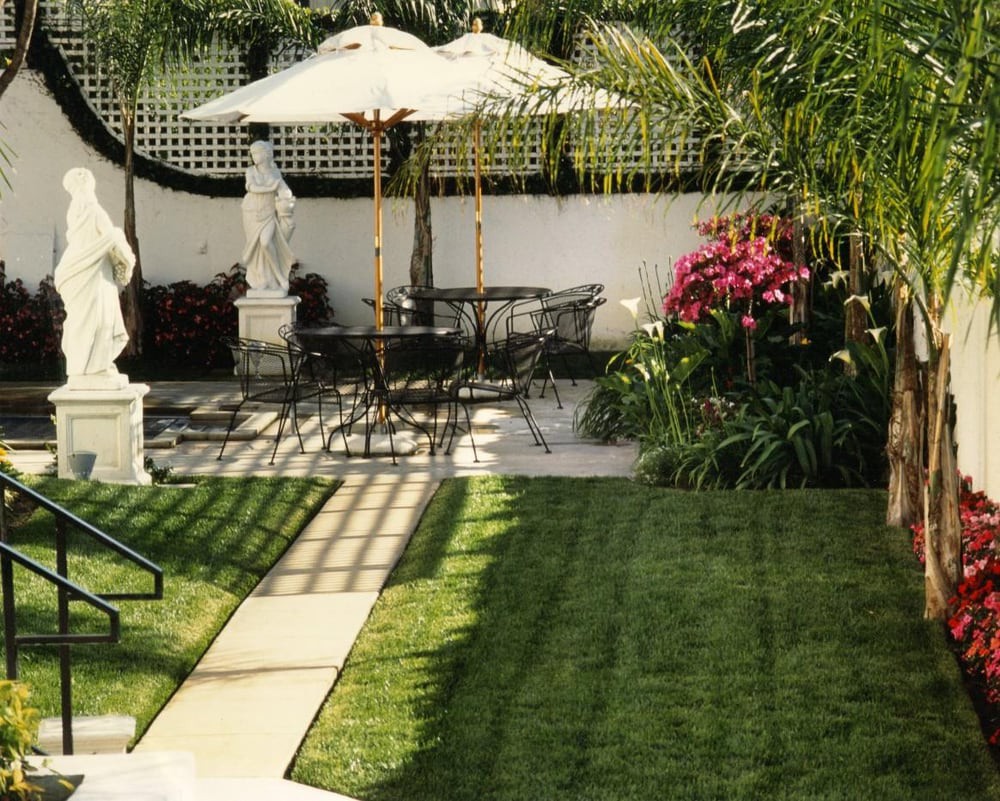 A tranquil garden patio with seating, an umbrella, statues, and lush greenery.