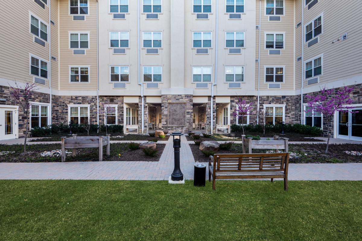 A view of an apartment building with a landscaped courtyard featuring benches and a stone facade.