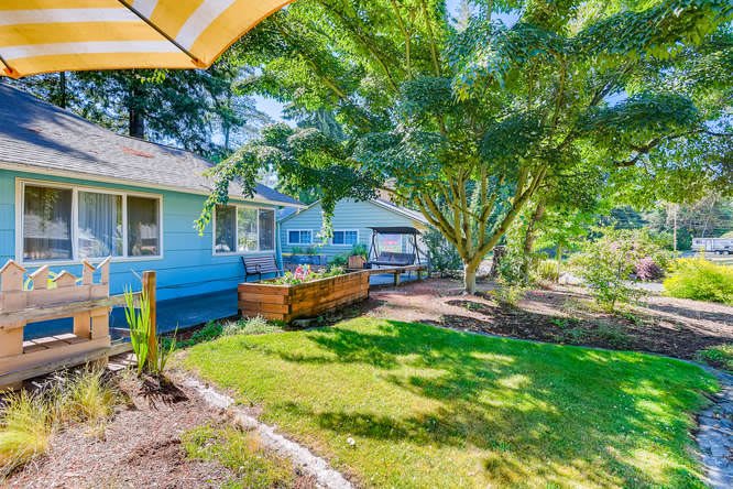 A sunny backyard featuring a well-kept garden and lush grass, surrounded by trees with a view of the house.