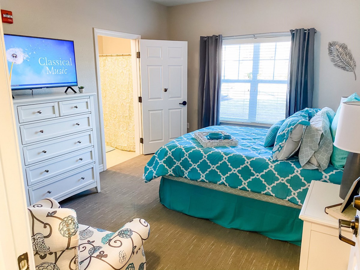 A cozy bedroom with turquoise bedding, a dresser with a TV, and a large window.