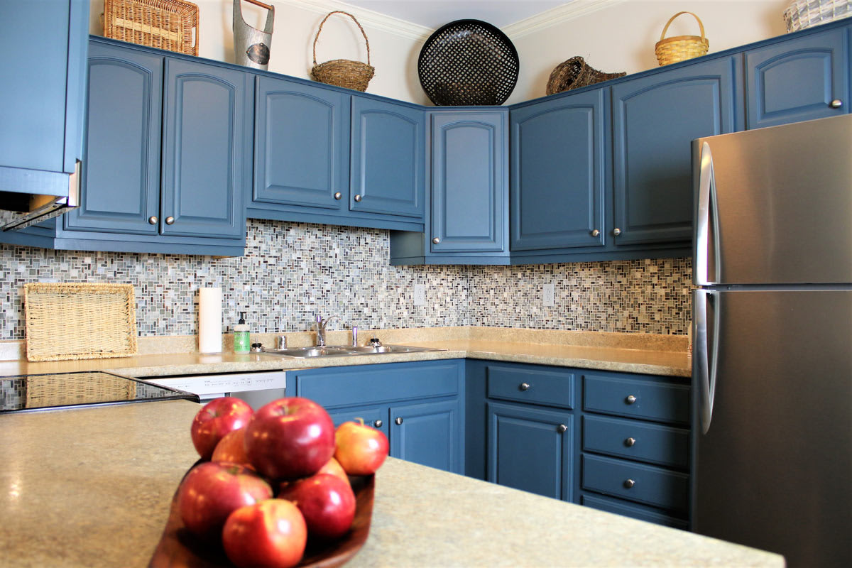 A cozy kitchen with blue cabinets, a fruit bowl filled with apples on the countertop, and a tiled backsplash.