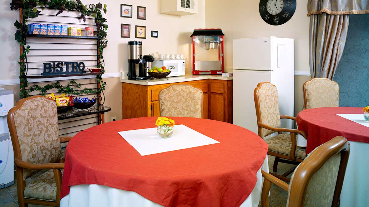 A cozy kitchen area featuring a dining table with a red tablecloth, surrounded by chairs, with appliances and decorative items in the background.