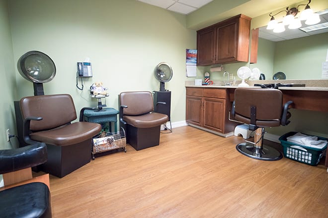 A cozy salon waiting area with chairs, a mirror, and hair dryers against green walls.