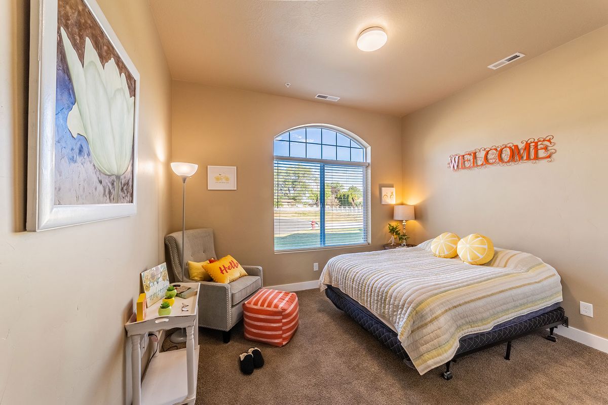 A cozy bedroom featuring a comfortable bed with yellow pillows, a reading chair with a lamp, and a large window providing natural light.