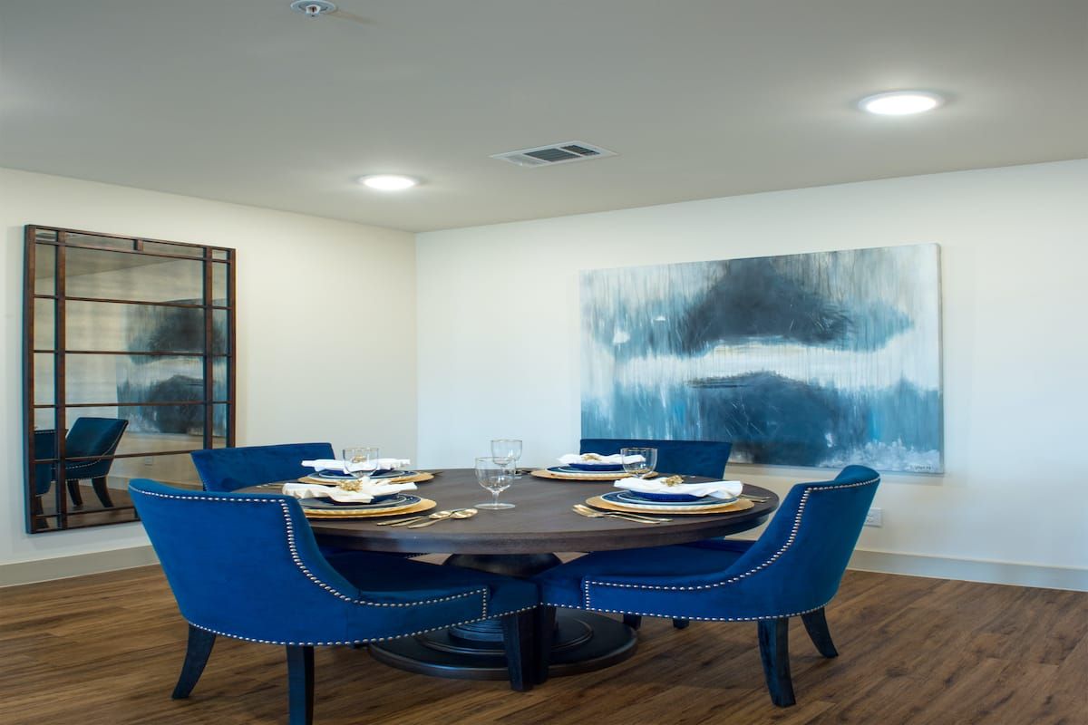 A modern dining room with a round table and blue upholstered chairs, featuring a large abstract painting on the wall.