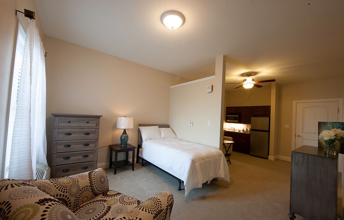 A cozy bedroom with a single bed, dresser, and chair, featuring neutral tones and warm lighting.