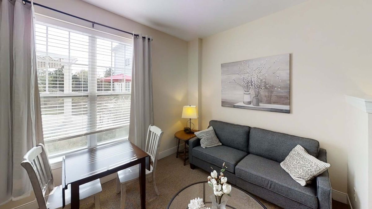 A cozy living room with a sofa, coffee and dining tables, a lamp, and wall artwork, lit by natural light from a large window.