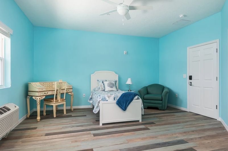 A minimalist bedroom with blue walls, featuring a single bed, wooden chair, armchair, and a wooden floor.