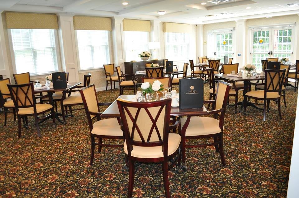 A well-decorated dining area with multiple tables and chairs, featuring elegant carpeting and large windows providing ample natural light.