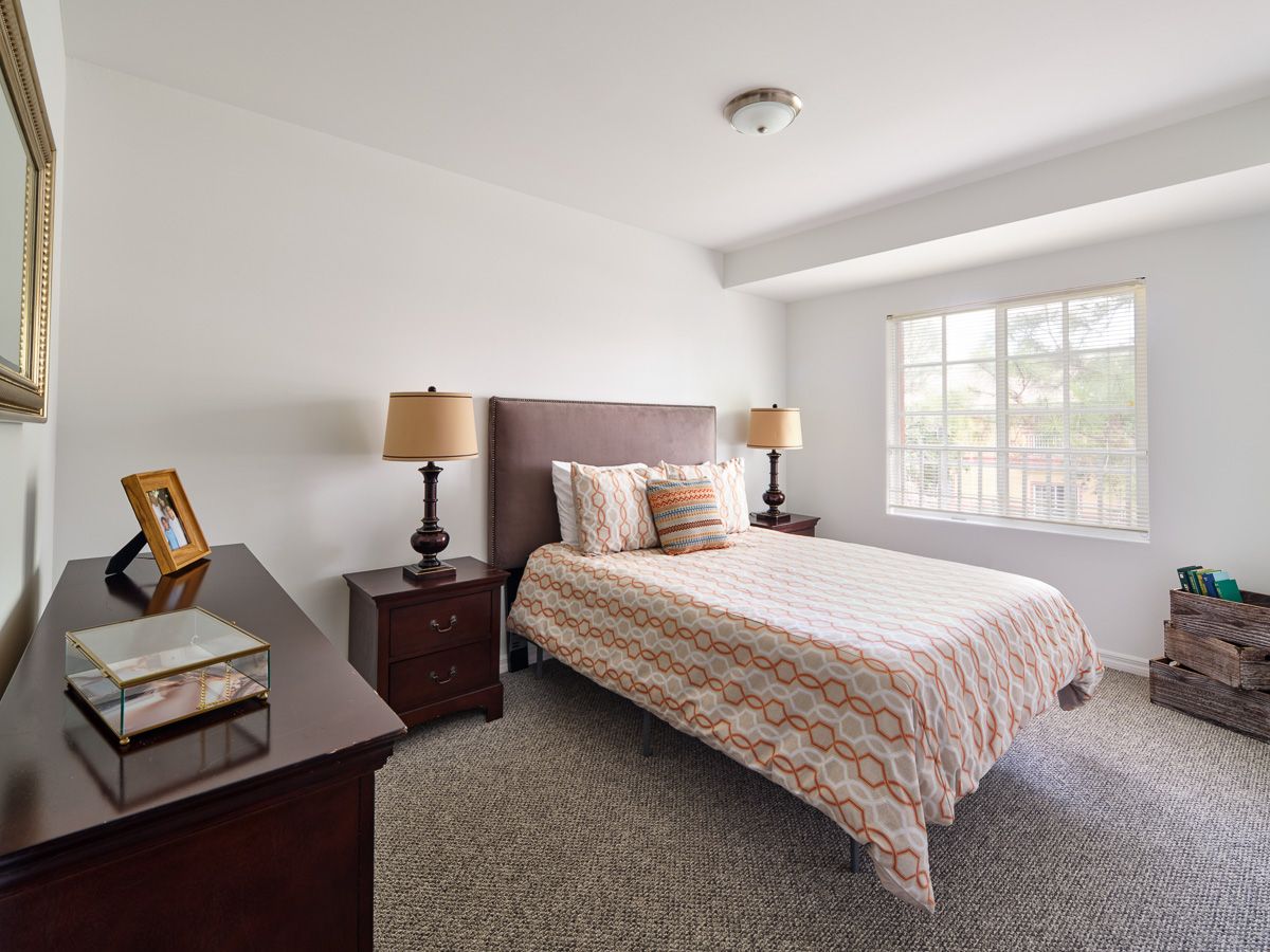 A bright bedroom with a neatly made bed, flanked by nightstands and lamps, featuring a window that lets in natural light.