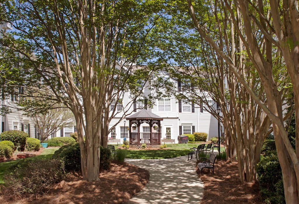 A scenic pathway lined with trees leads to a residential building, complemented by well-maintained landscaping and a bench for relaxation.