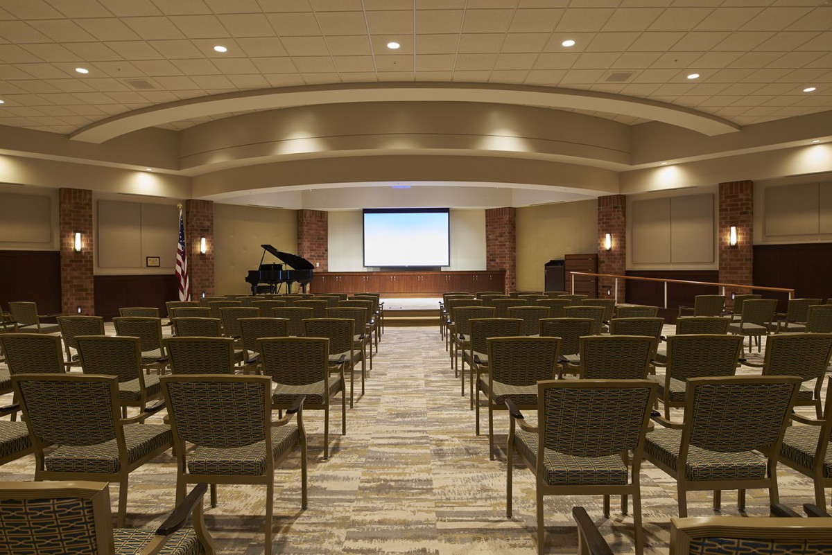 A spacious auditorium with rows of chairs facing a stage equipped with a grand piano and a projector screen.