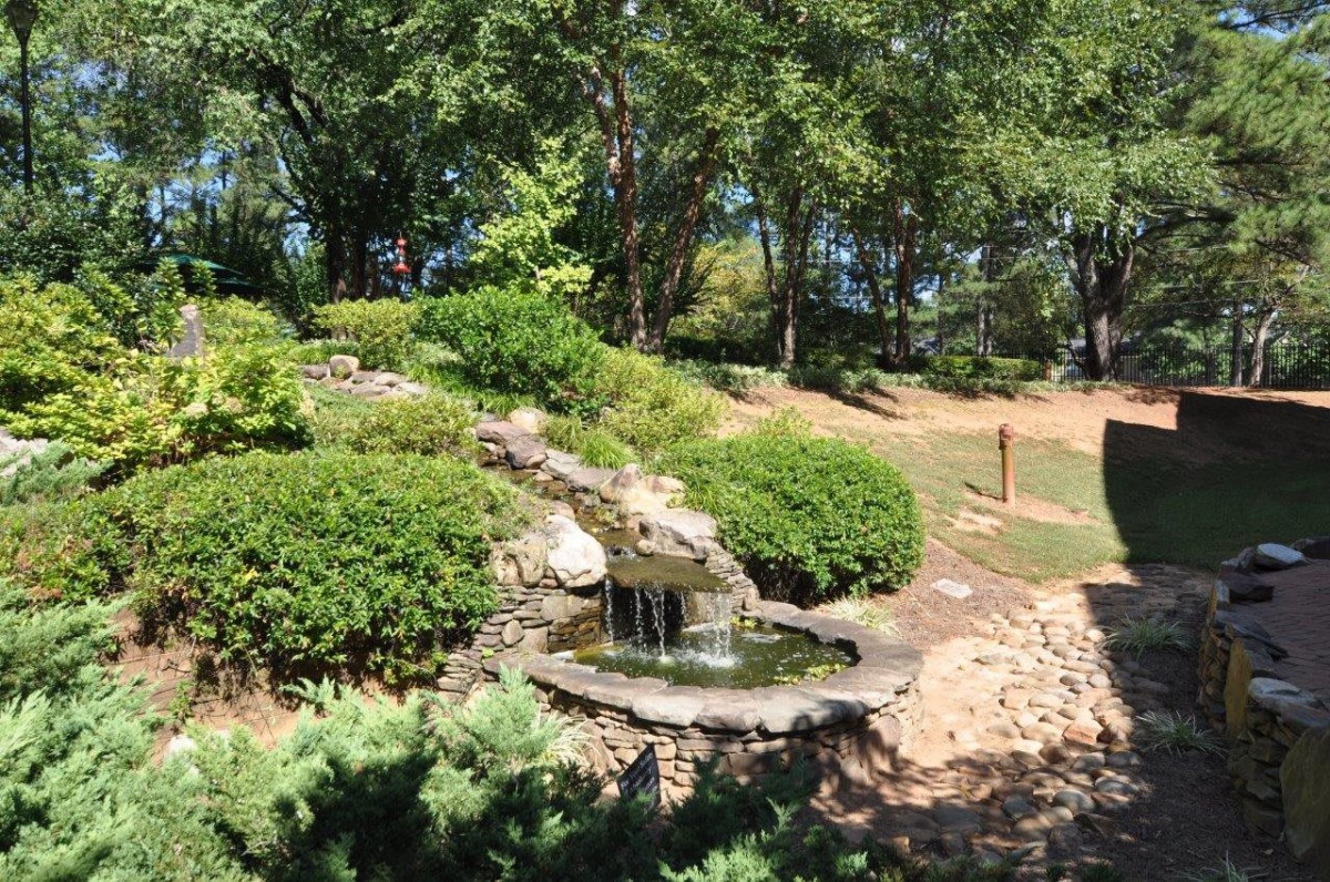 A serene garden landscape featuring a small waterfall flowing into a pond, surrounded by lush greenery and trees.