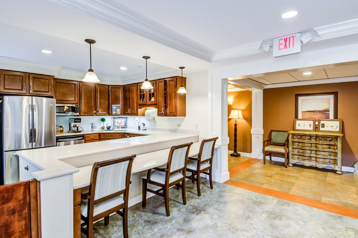 A modern kitchen with wooden cabinets and a long dining countertop equipped with several chairs. Pendant lights illuminate the space, offering a cozy atmosphere.