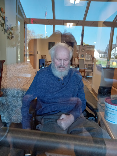 An elderly man sitting in a wheelchair, lost in thought as sunlight filters through the window.