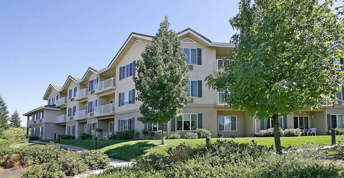 A residential apartment building surrounded by trees and well-maintained landscaping on a sunny day.