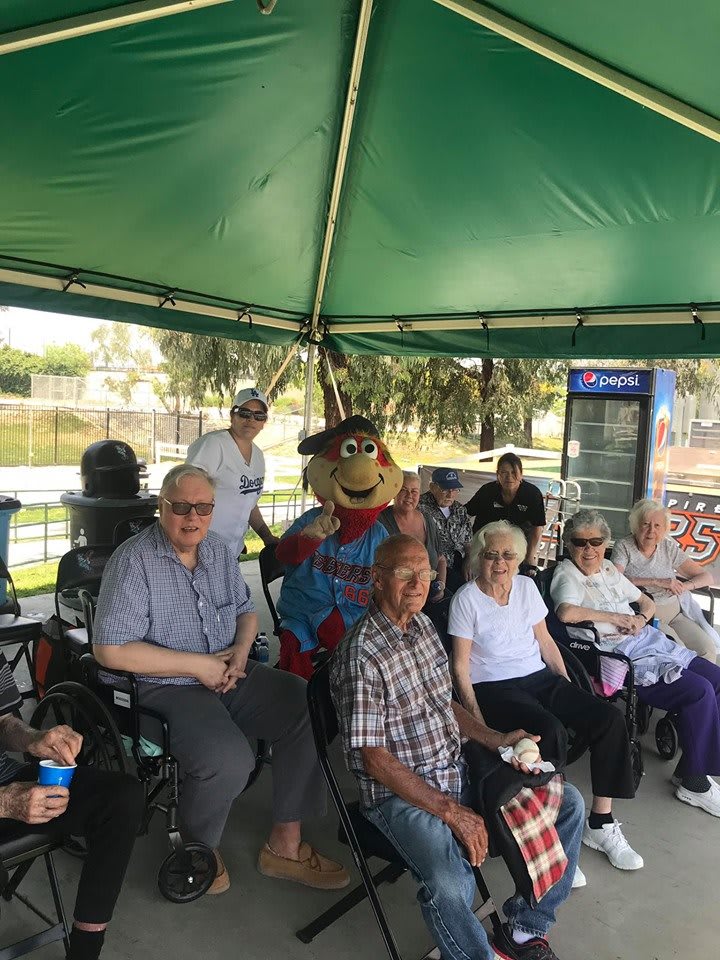 A group of elderly individuals sitting under a green tent, accompanied by someone in a colorful costume.
