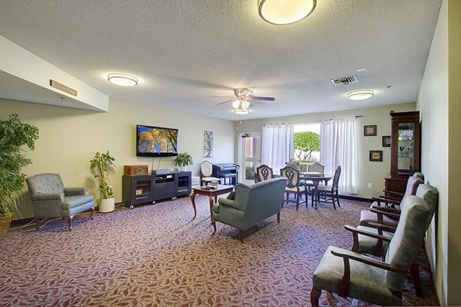A cozy living room with a television, comfortable seating, and natural light from large windows.