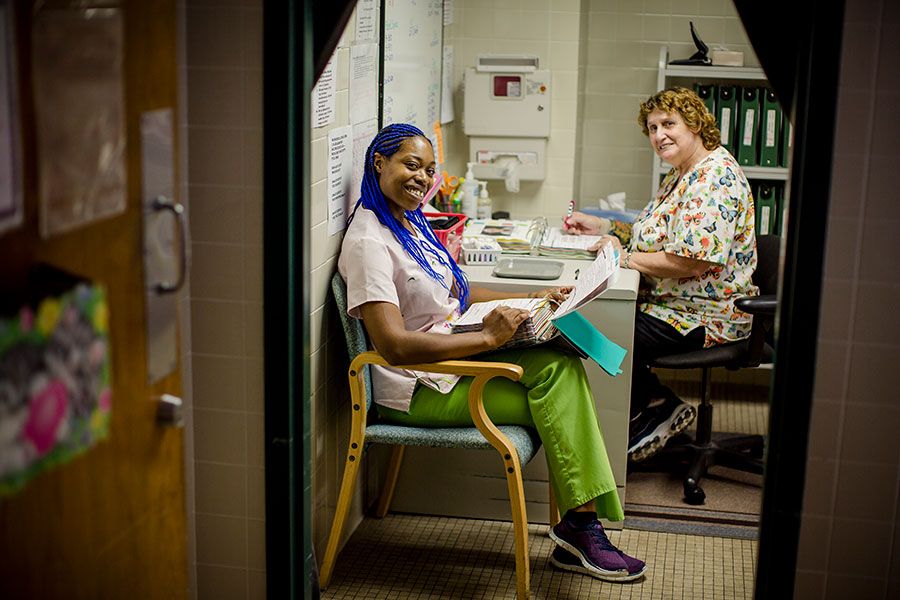 Two people smiling and working in an office setting, one sitting at a desk and the other on a chair.