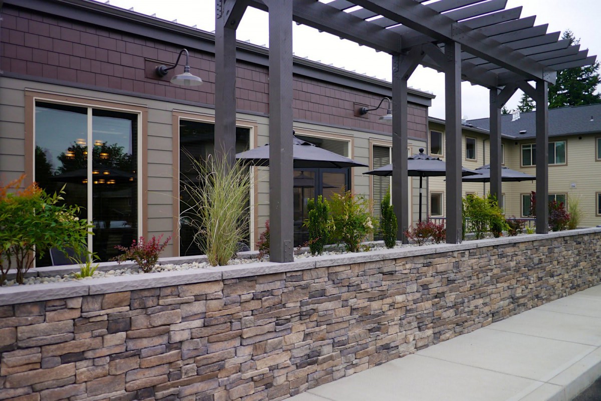 A modern building with a stone wall and landscaped exterior, featuring a pergola and decorative plants.