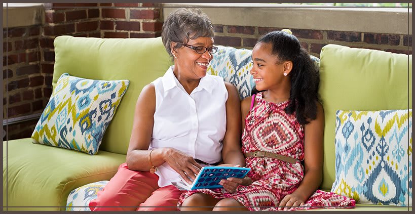 A grandmother and granddaughter sitting on a couch, smiling while looking at a tablet together.
