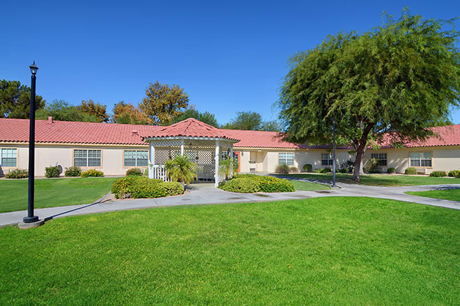 A sunny day in a suburban neighborhood, featuring a residential building with a lush green lawn and trees.