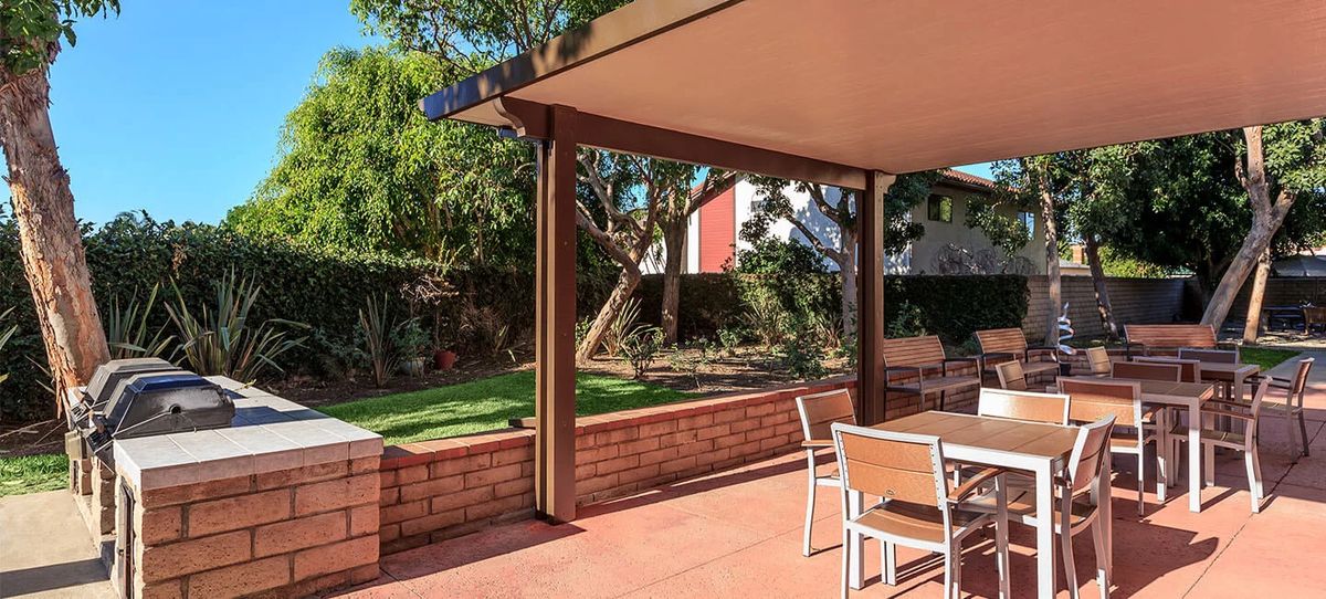 A sunny backyard patio with outdoor seating, surrounded by a garden and trees.