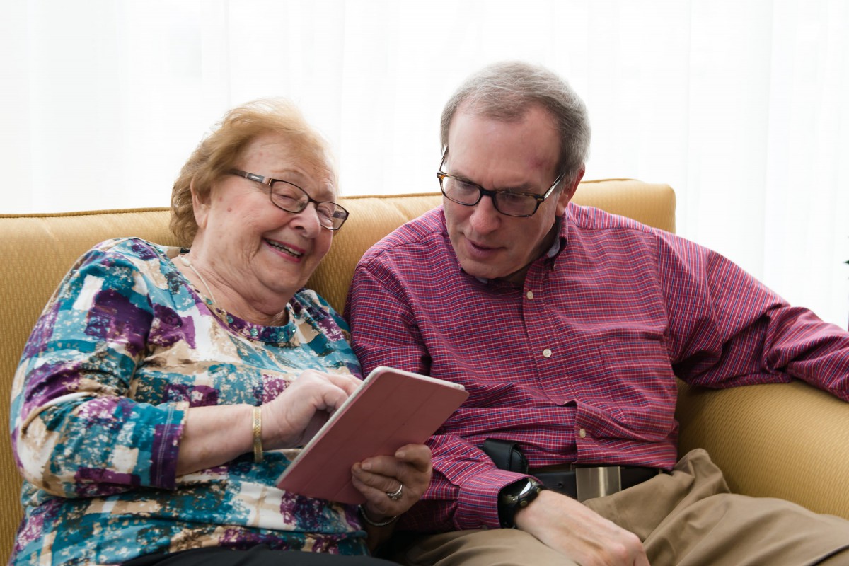 An elderly woman and a middle-aged man sit together on a couch, smiling as they look at a tablet.